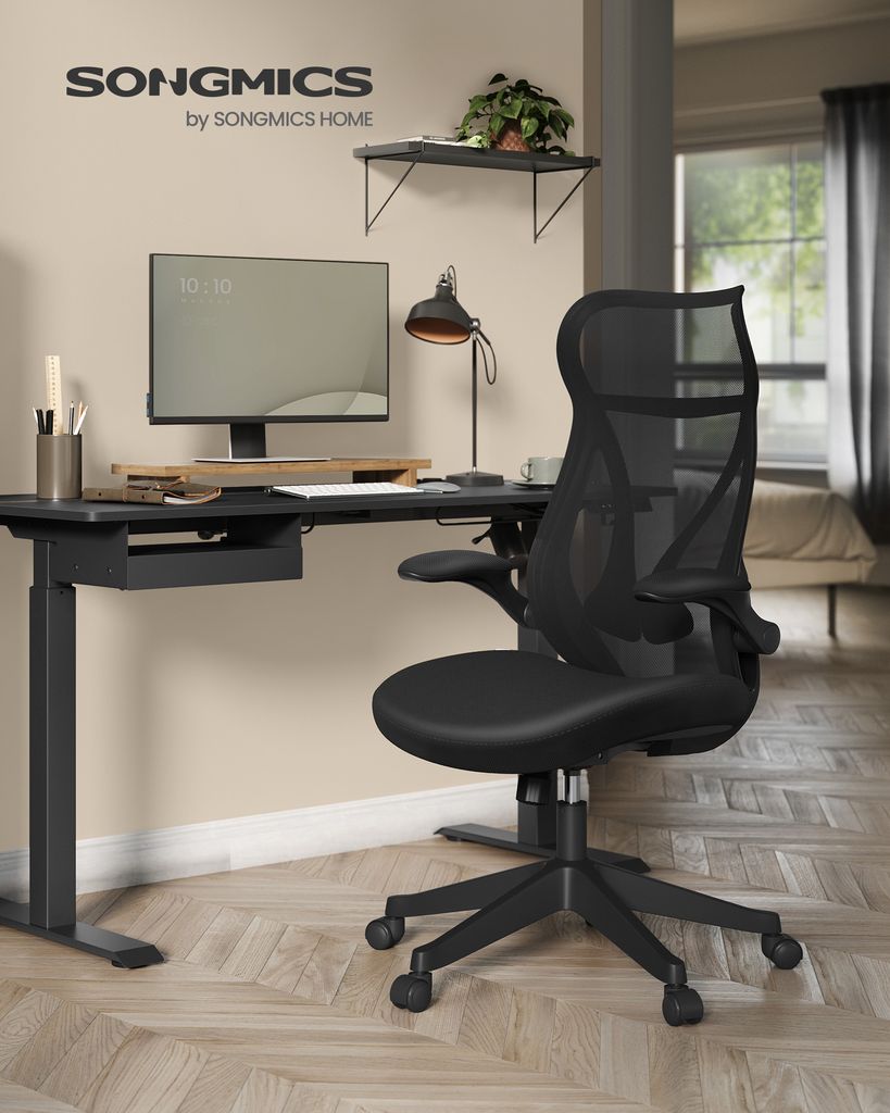 A black ergonomic office chair is placed next to a black standing desk with a computer monitor, lamp, and desk accessories.