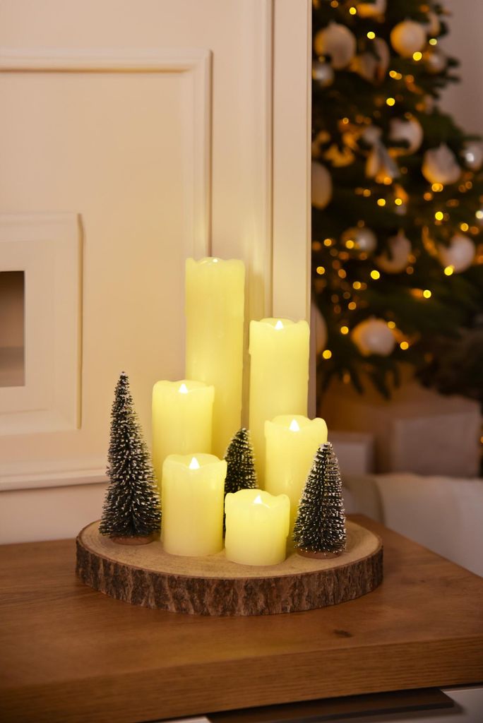 A Christmas-themed centerpiece with LED candles and miniature trees on a wooden base, with a decorated Christmas tree in the background.