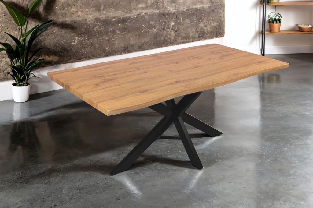 A wooden dining table with a black spider leg base is shown in a room with a concrete wall and a plant.