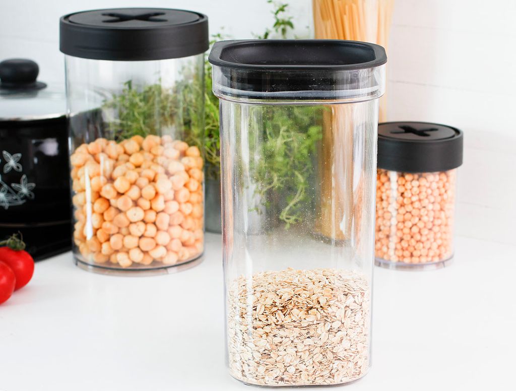 Three clear plastic food storage containers with black lids are arranged on a white surface. The containers hold different dry goods: chickpeas, oats, and lentils. In the background, there are some herbs and a black pot.