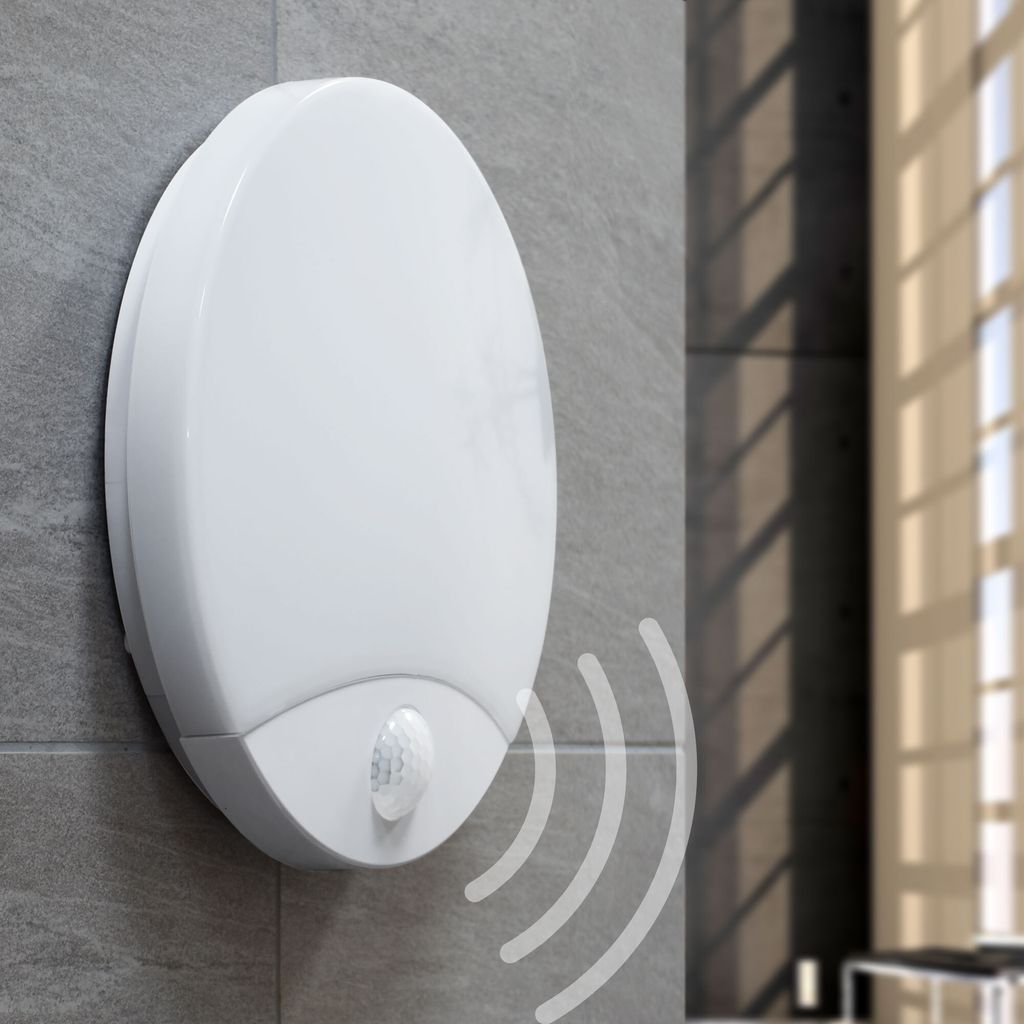 A white motion-sensing outdoor light fixture mounted on a gray tiled wall, with sunlight casting shadows through a window in the background.