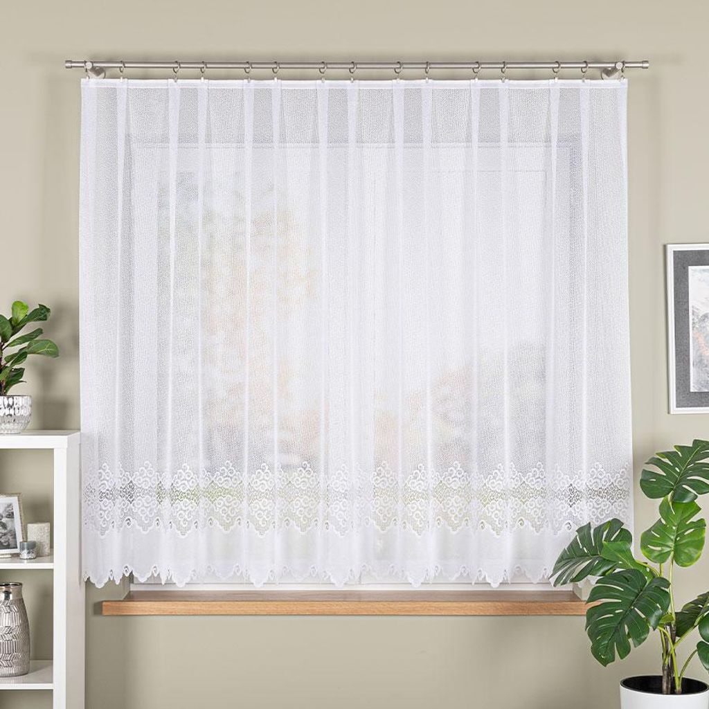 A white lace curtain hangs in a window, with a wooden windowsill visible below. A white shelf unit with decorative items is to the left, and a potted plant with large leaves is to the right.