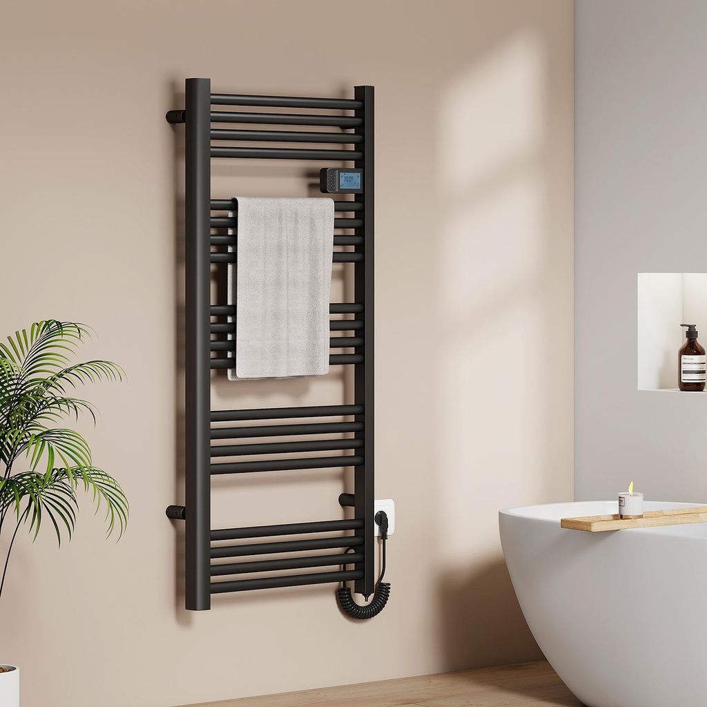 A black electric towel warmer is mounted on a light beige wall in a bathroom, with a white towel draped over it. A plant is visible to the left, and a white bathtub with a wooden tray and a candle is to the right.
