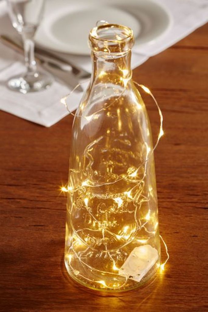 A clear glass bottle filled with white LED string lights sits on a wooden table.