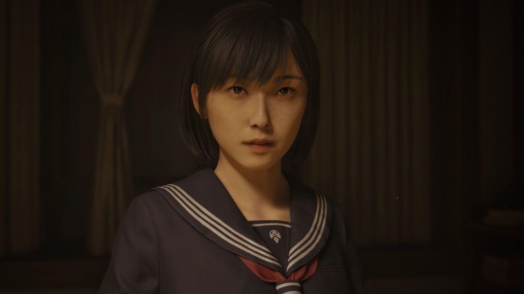 A young woman in a school uniform looks directly at the camera in a dimly lit room.