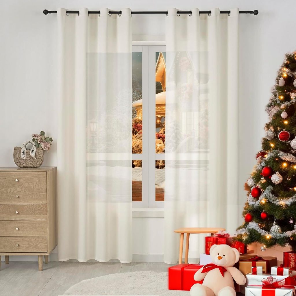 A decorated Christmas tree and gifts are placed next to a window with cream-colored curtains.