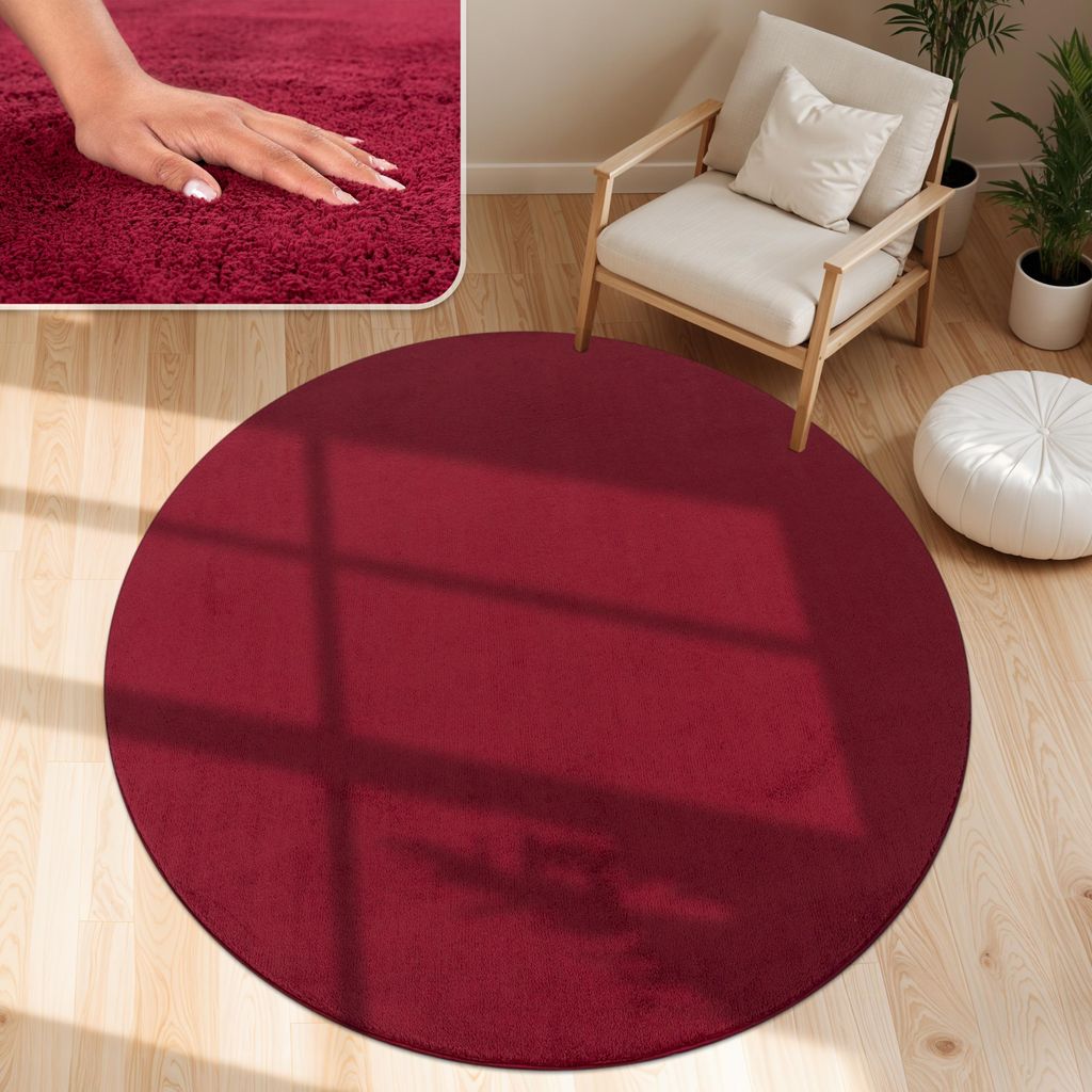 A round, red rug is placed on a wooden floor in a living room setting, with a chair and plants in the background.
