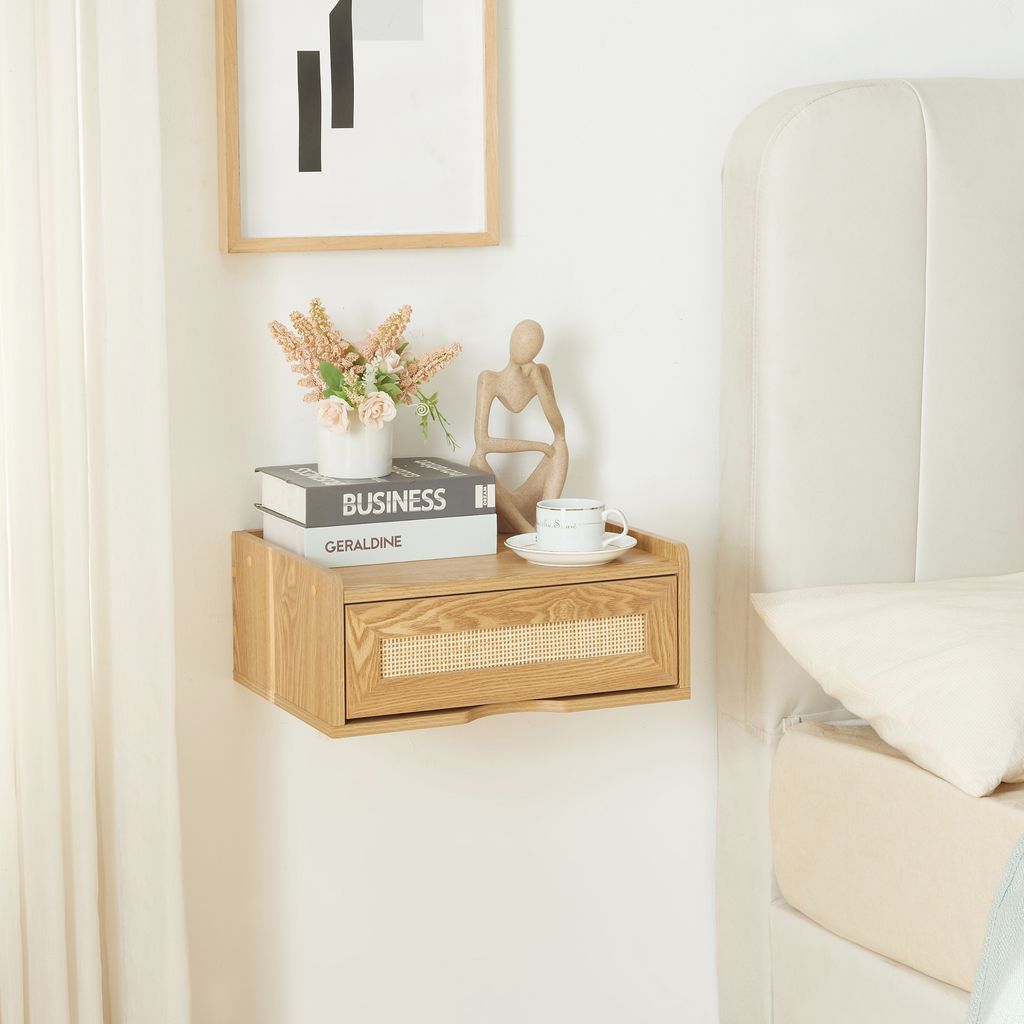 A wooden floating nightstand with a cane drawer front is mounted on a white wall next to a bed. It is decorated with books, a vase of flowers, and a decorative figurine.