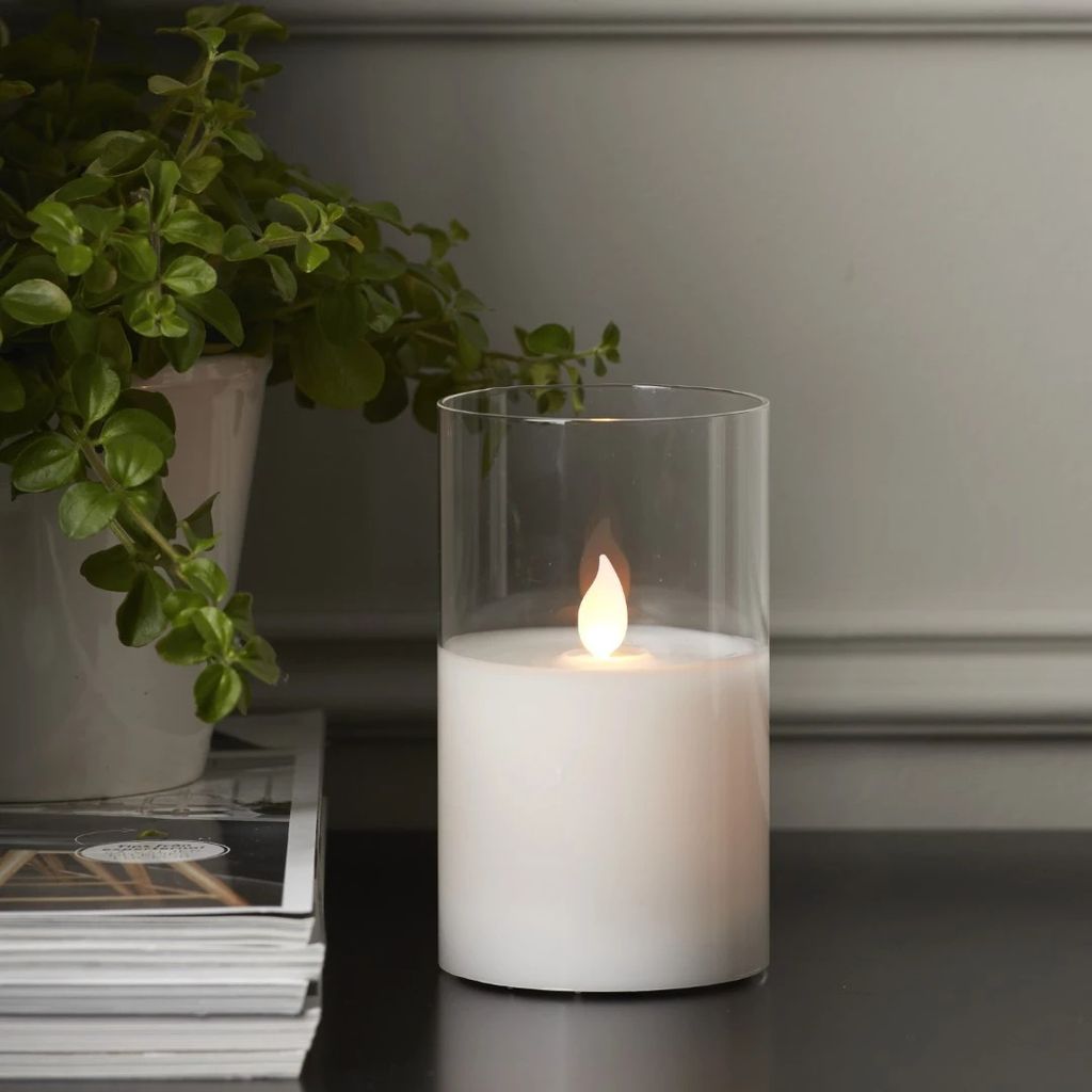 A white LED candle flickers inside a glass cylinder, placed on a dark surface next to a stack of magazines and a potted plant.