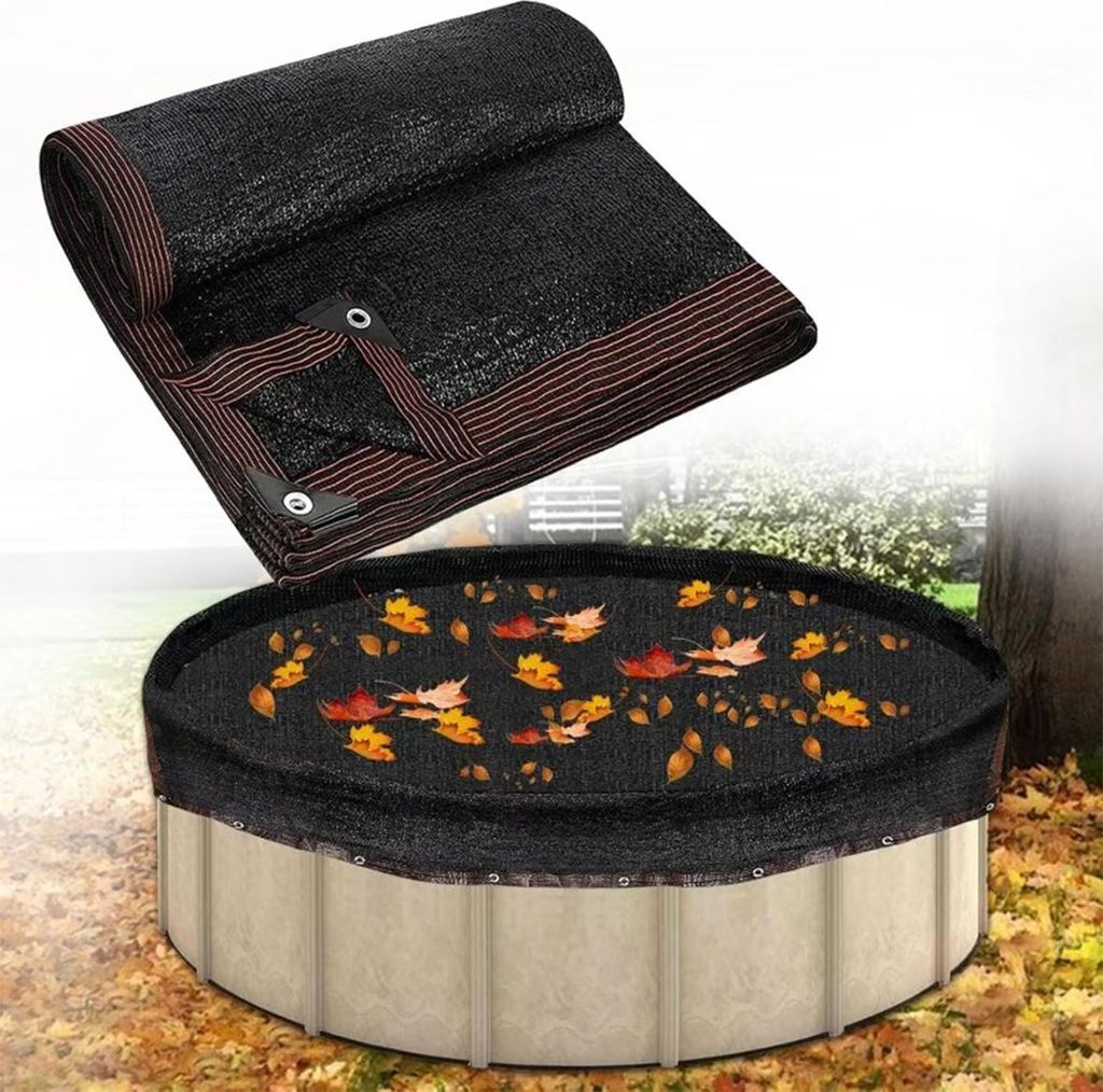 A black pool cover is shown on top of an above-ground pool, with fallen leaves scattered on the cover. A rolled-up portion of the cover is also visible in the upper left corner.