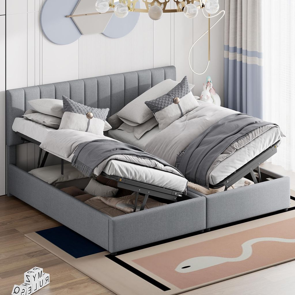 A bedroom setting with a grey upholstered bed frame, featuring two lift-up storage compartments. The bed is made with white and grey bedding and pillows. A decorative wall art and curtains are visible in the background.