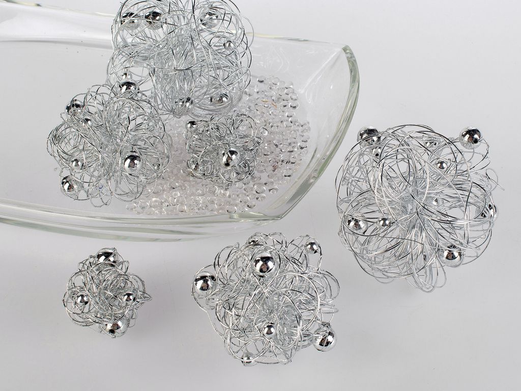 Several decorative wire balls with silver beads are arranged on a clear glass dish filled with small clear beads, with some balls placed outside the dish on a light gray surface.