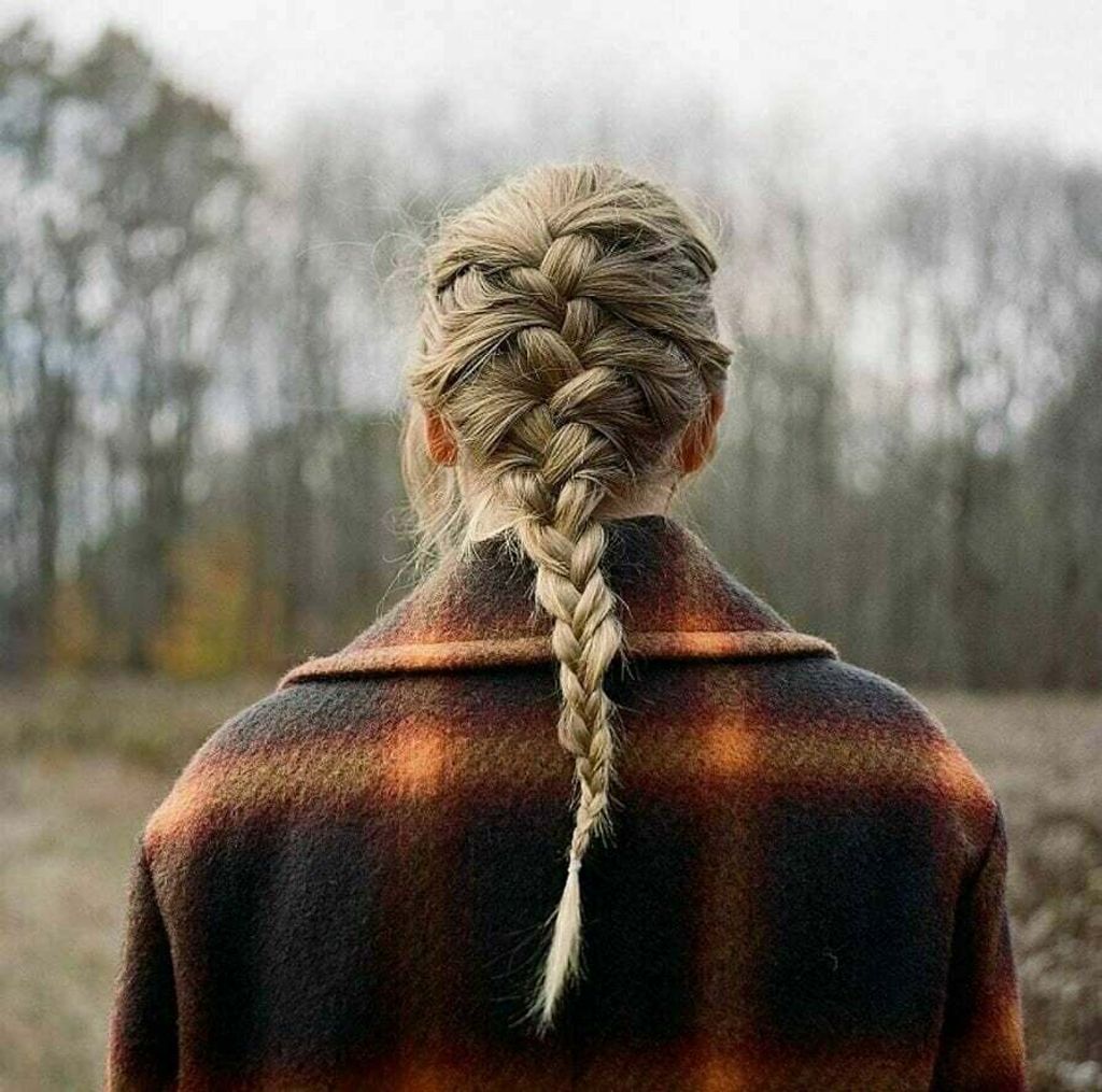 A person from behind wearing a plaid coat and a braided hairstyle, standing in an outdoor setting with trees in the background.