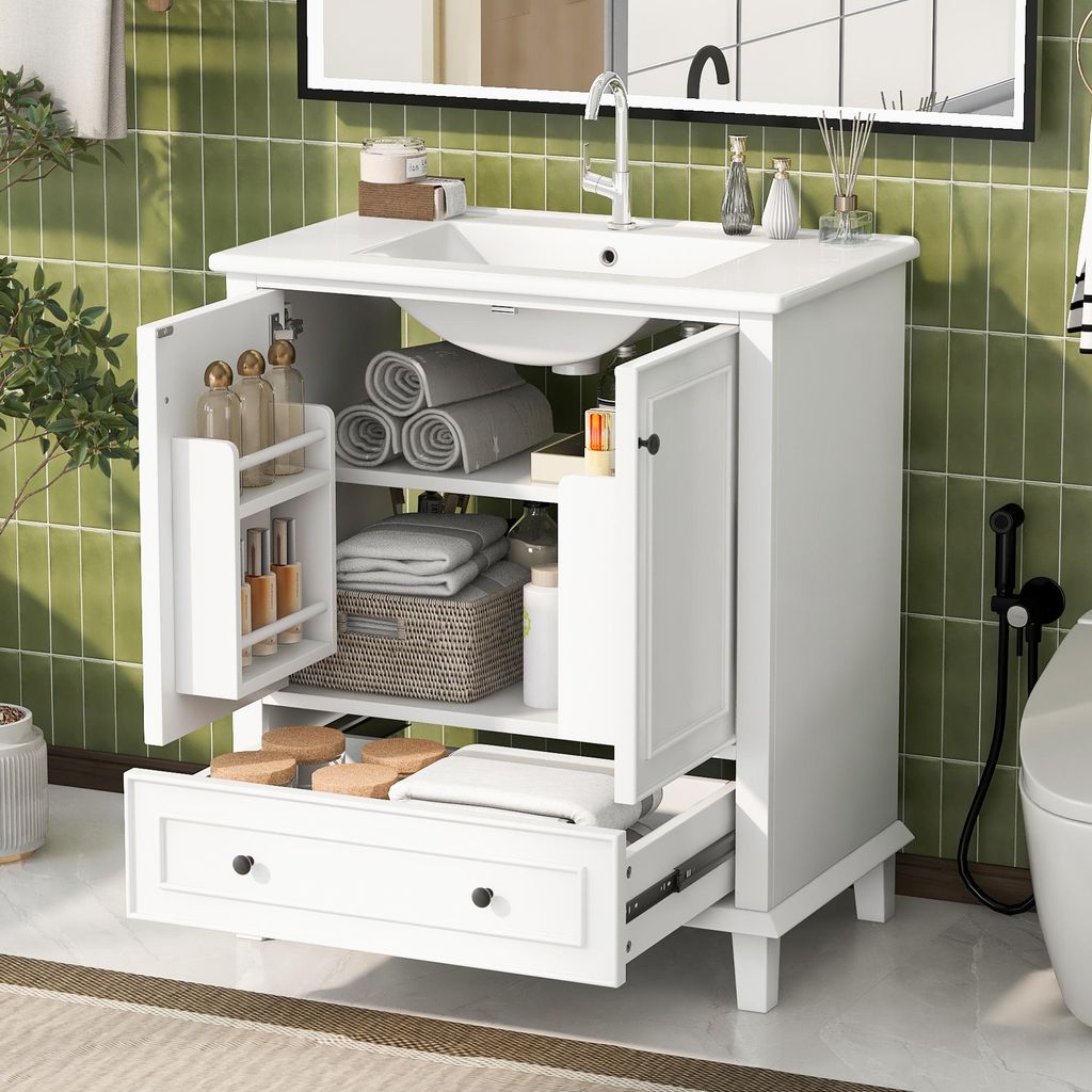 A white bathroom vanity with its doors and drawer open, revealing storage shelves with towels, toiletries, and a basket. The vanity is set against a green tiled wall with a mirror above it.