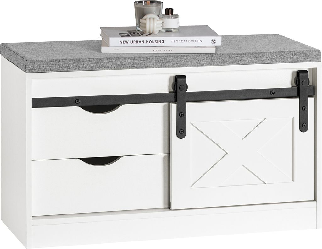 A white shoe bench with a gray upholstered top, featuring two drawers and a sliding barn door with a decorative 'X' pattern. Books and decorative items are placed on top.