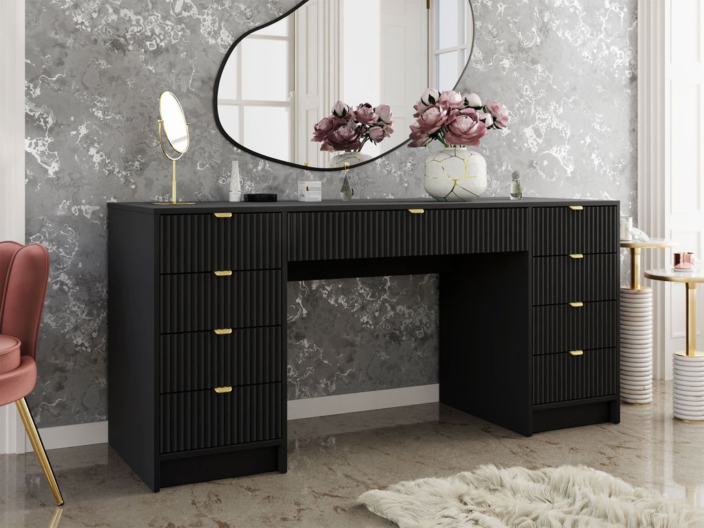 A black vanity table with gold handles and drawers, set in a room with a decorative gray and white wallpaper and a pink chair.