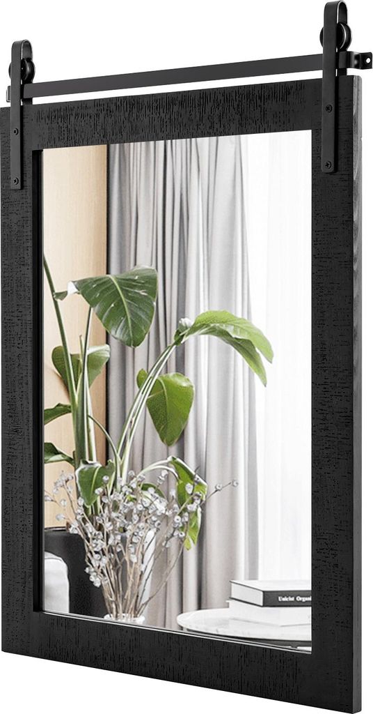 A black wooden framed mirror with a sliding barn door mechanism is shown. Inside the mirror, a plant in a vase and a stack of books are reflected.