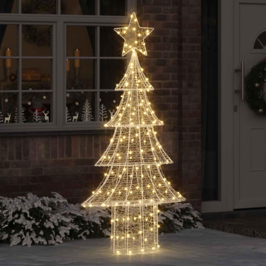 A lighted Christmas tree decoration placed outdoors on a snowy ground, next to a brick wall and a window.