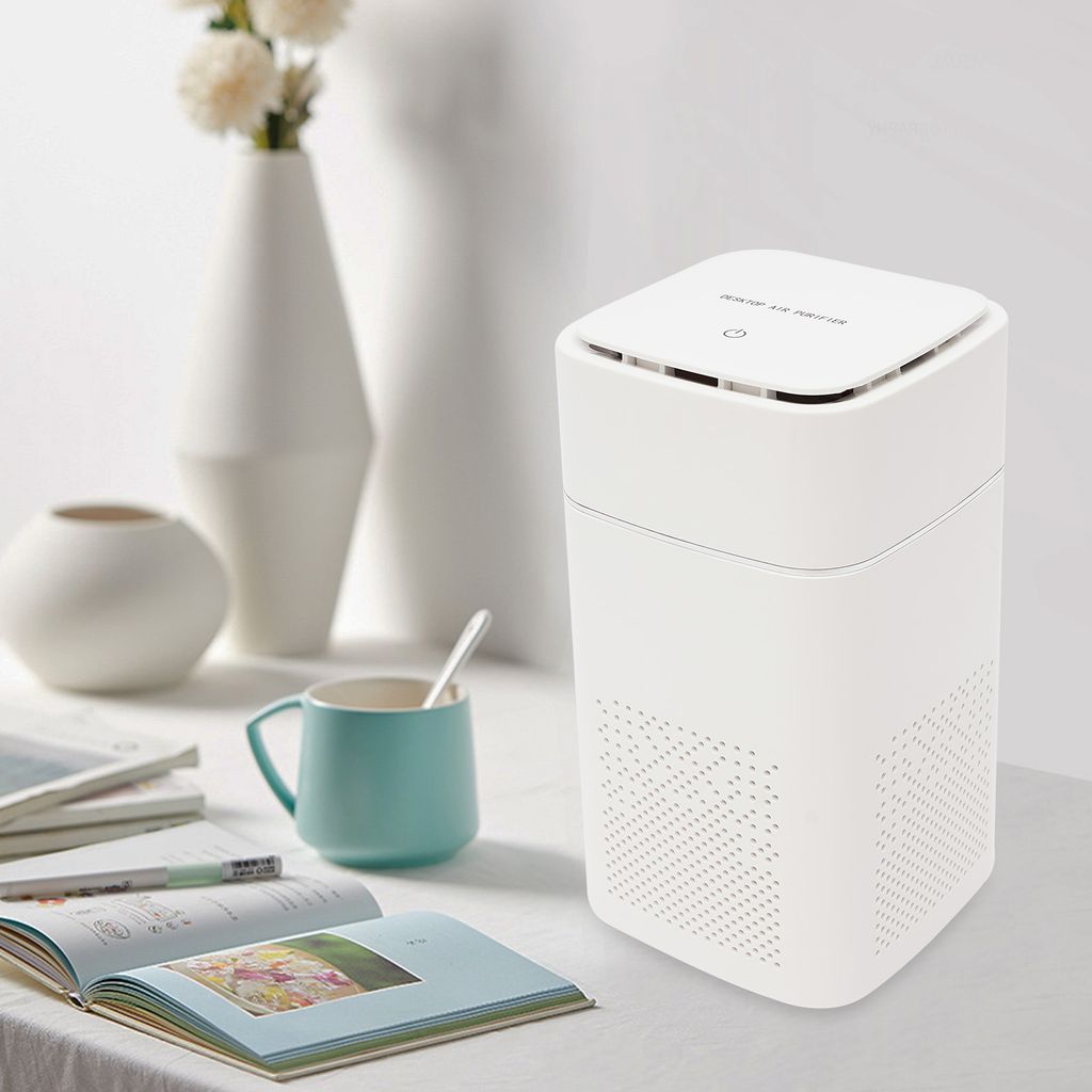 A white air purifier sits on a table next to a mug and some books, with vases in the background.