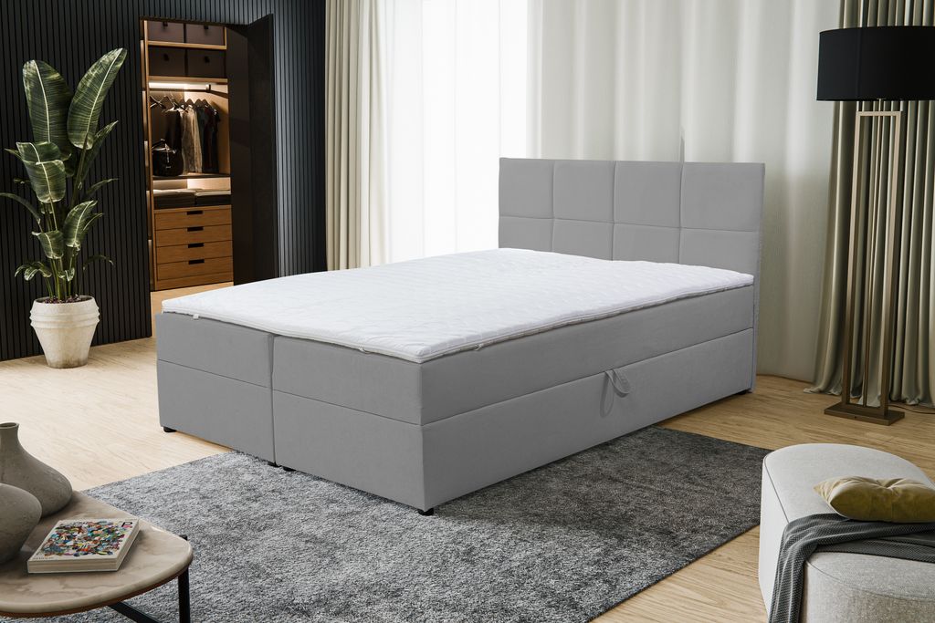 A bedroom scene featuring a gray boxspring bed with a white mattress and headboard. A large plant is visible in the background, along with a closet and a floor lamp.