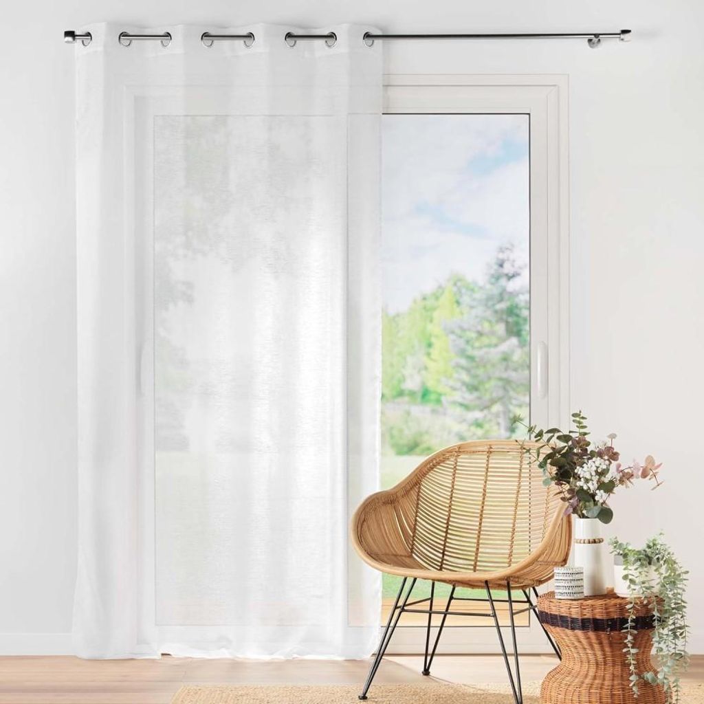 A white sheer curtain is partially drawn across a window, with a wicker chair and a small table with flowers next to it.