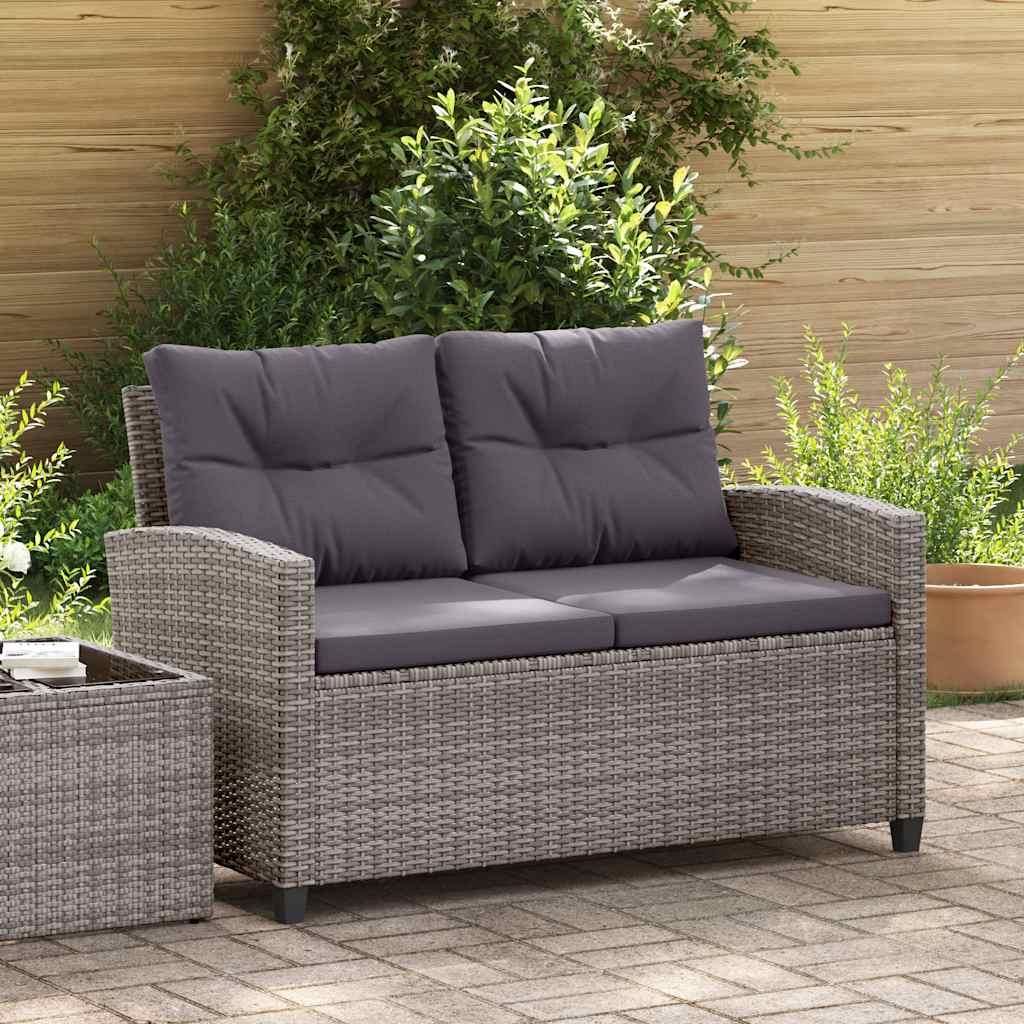 A grey rattan two-seater sofa with dark grey cushions is placed on a paved patio, surrounded by greenery and a wooden fence.
