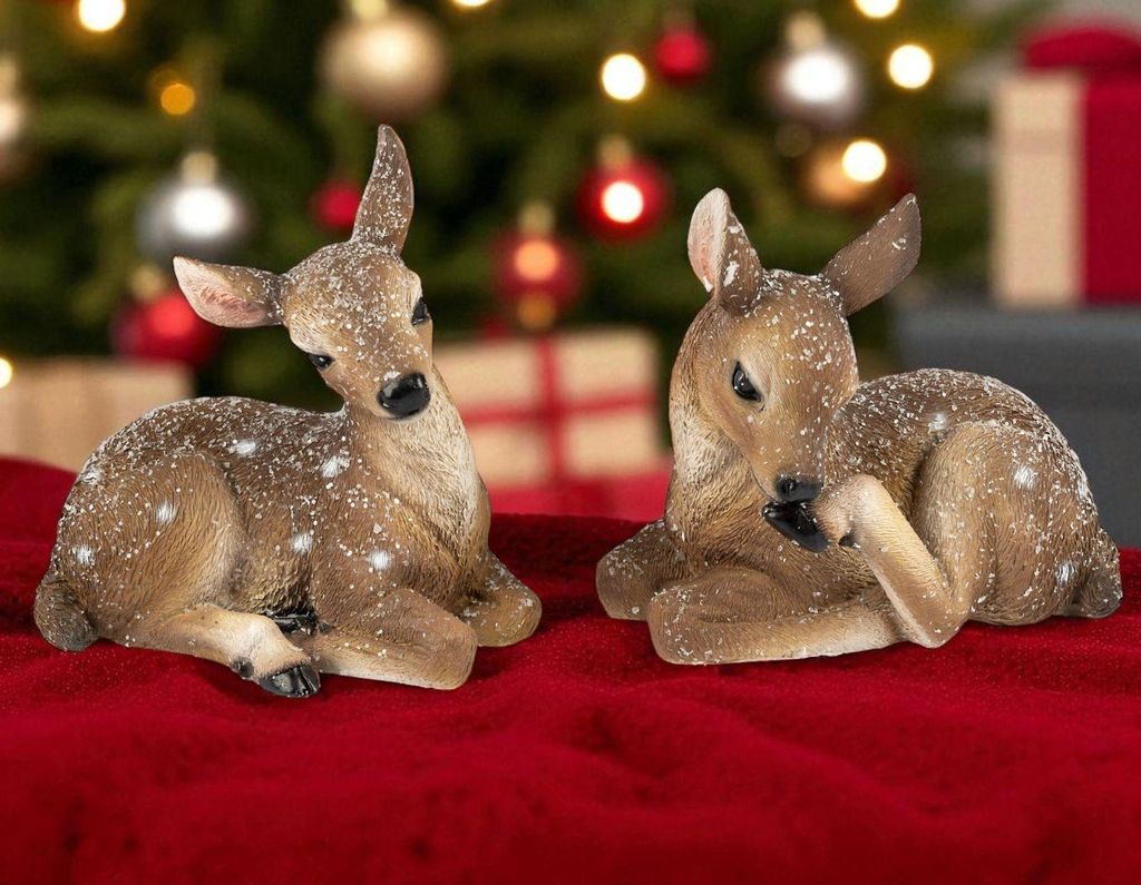 Two fawn figurines are lying down on a red surface, with a blurred Christmas tree and gifts in the background.