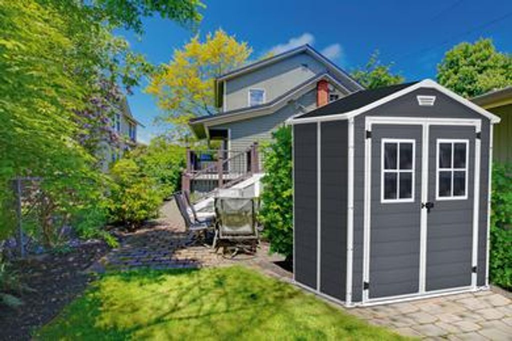 A dark gray Keter garden shed is shown in a backyard setting, surrounded by lush green trees and grass, with a house and patio in the background.