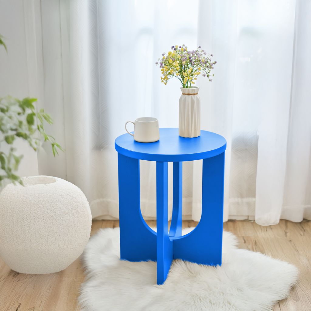 A blue side table with a mug and a vase of flowers on it, placed on a white rug in a room with sheer curtains.