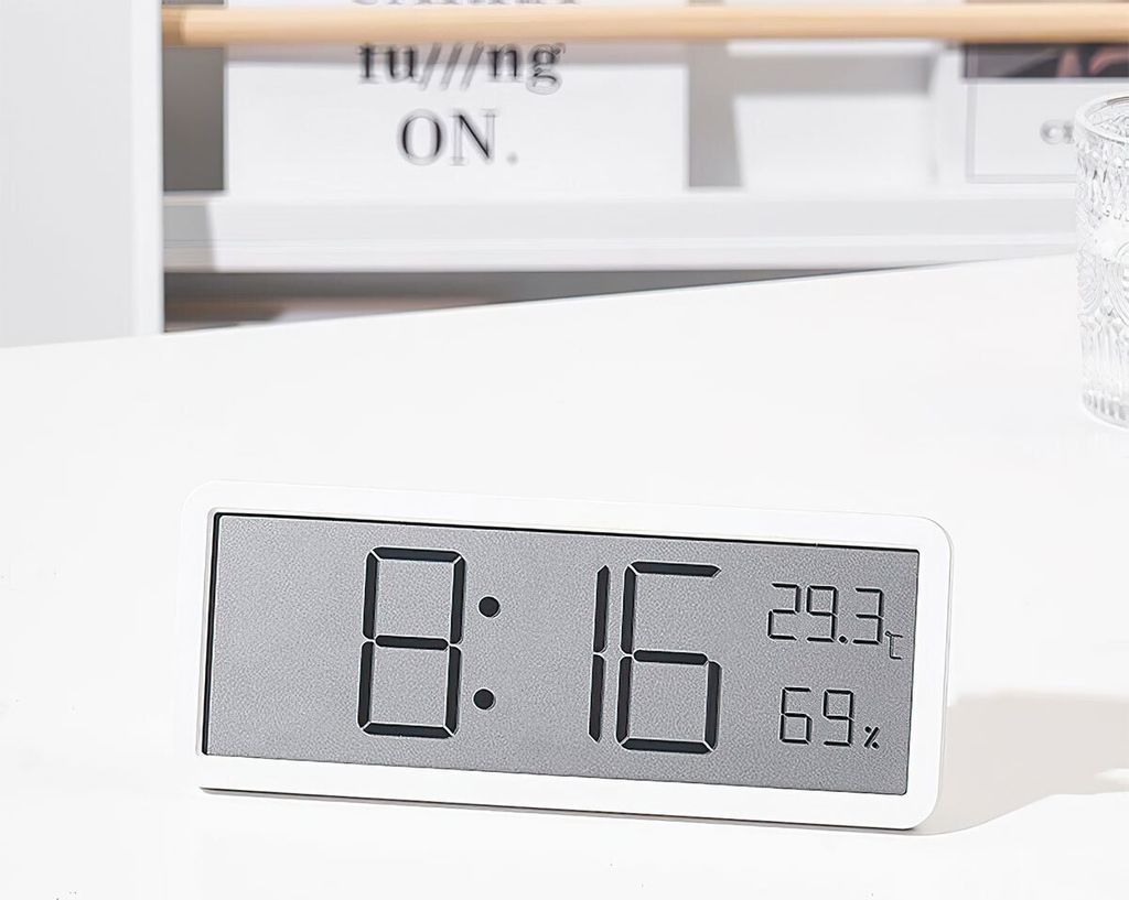 A digital clock displaying time, temperature, and humidity on a white desk.