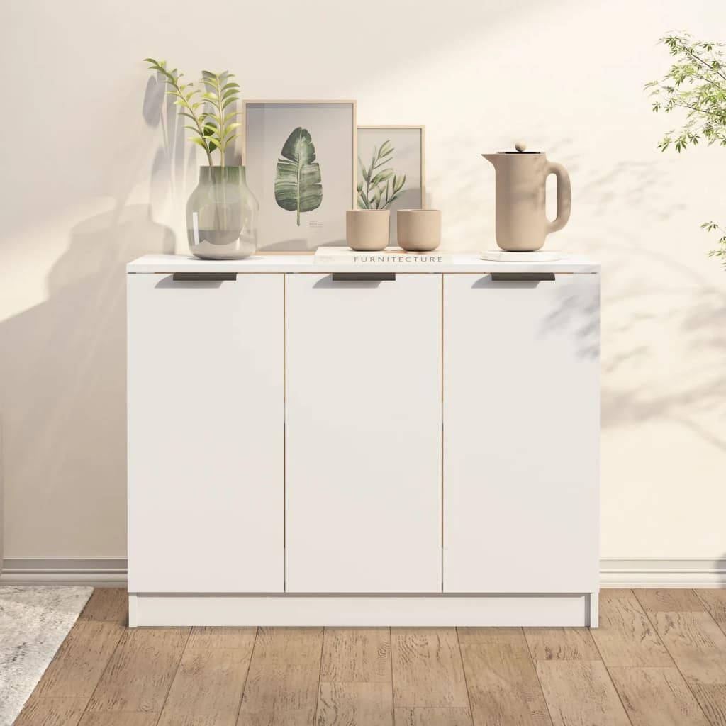 A white sideboard with three doors is shown against a light-colored wall. The top of the sideboard is decorated with a vase of plants, two small cups, and a pitcher. The floor is made of wooden planks.
