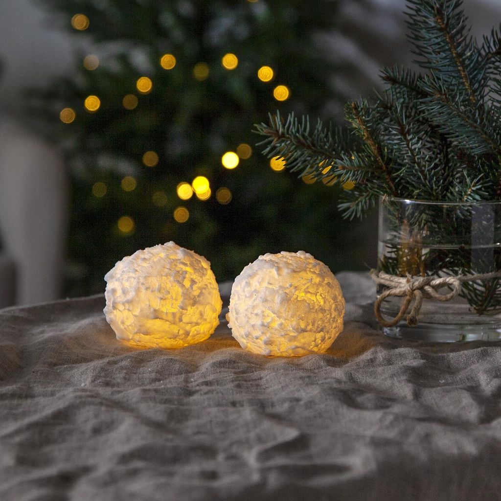 Two snowball-shaped candles are placed on a textured gray tablecloth, with a blurred Christmas tree and lights in the background.