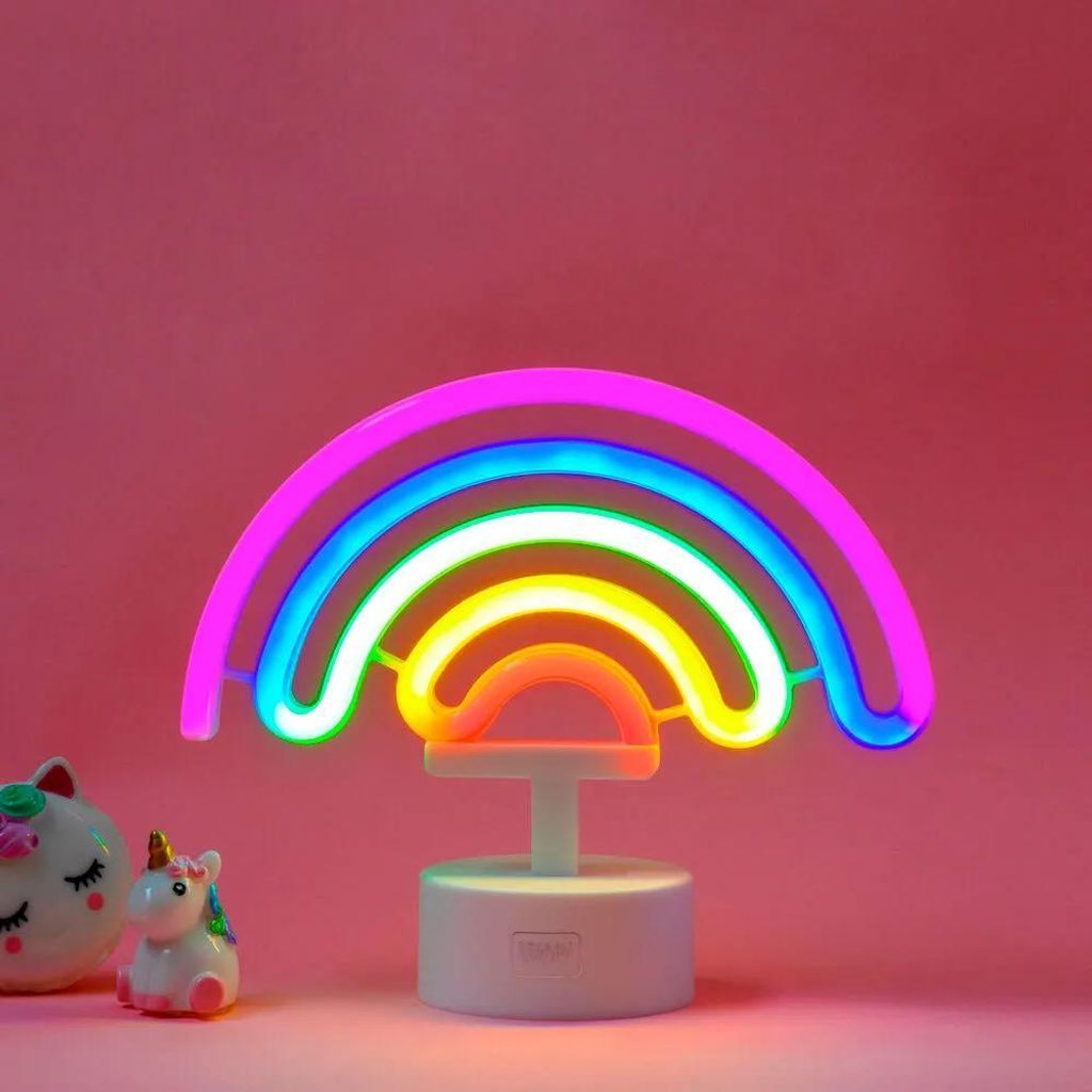 A neon rainbow lamp with a white base, placed on a pink background. Two small decorative unicorn figures are visible to the left of the lamp.