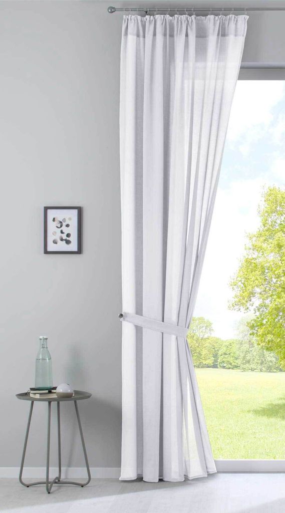 A sheer white curtain with gray stripes is tied back with a fabric band, hanging in front of a window with a view of a green lawn and trees. A small side table with a bottle and a framed picture on the wall are also visible.