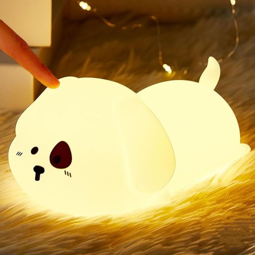 A finger is touching a cute, glowing puppy-shaped night light that is lying on a fluffy rug. Fairy lights are visible in the background.