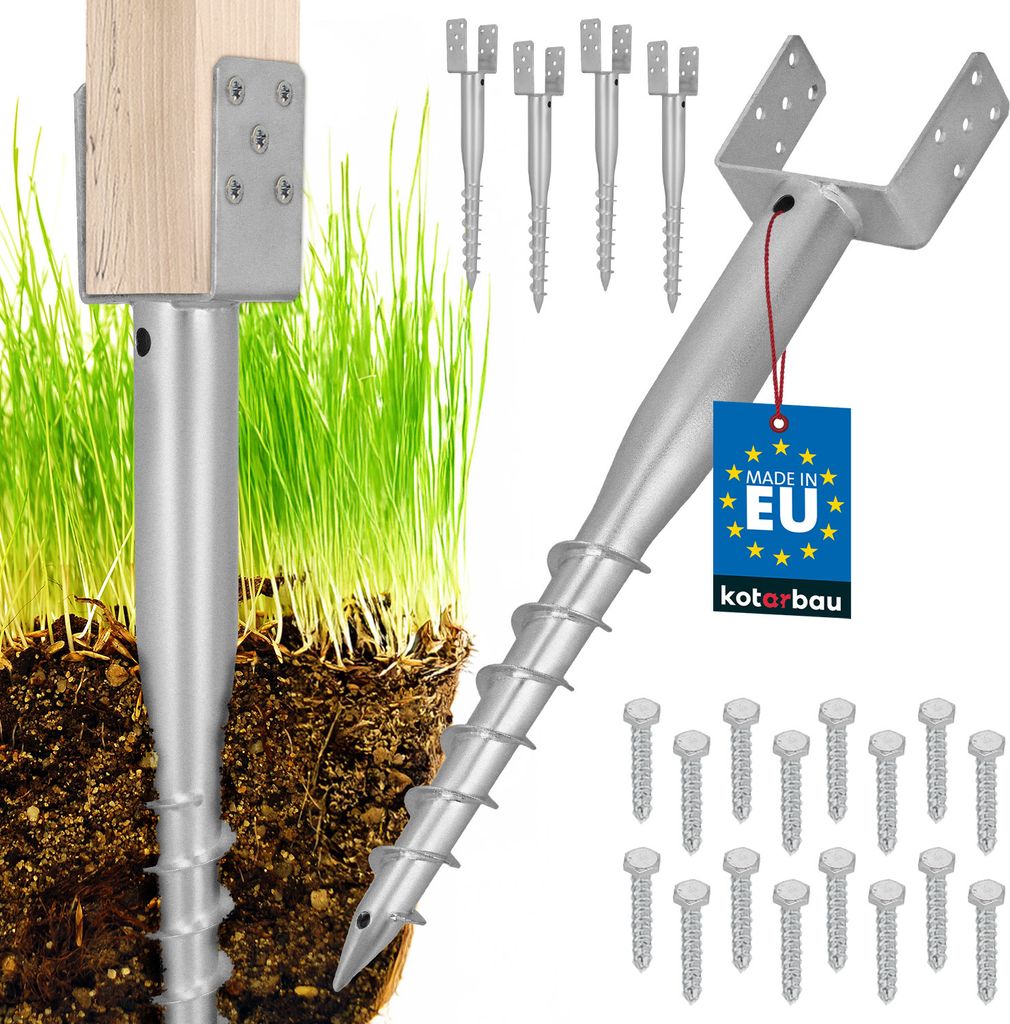 A close-up of metal ground anchors with a wooden post attached, set against a backdrop of green grass and soil. Several other anchors and screws are also visible.