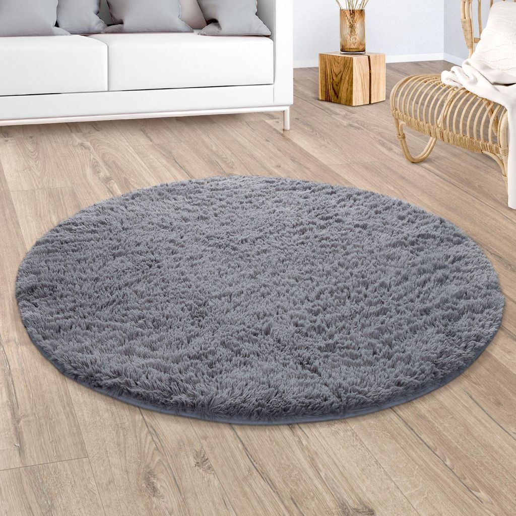 A round, fluffy gray rug is placed on a wooden floor in a living room setting. A white sofa is visible on the left, and a wicker chair is on the right. A small wooden side table with a vase is in the background.