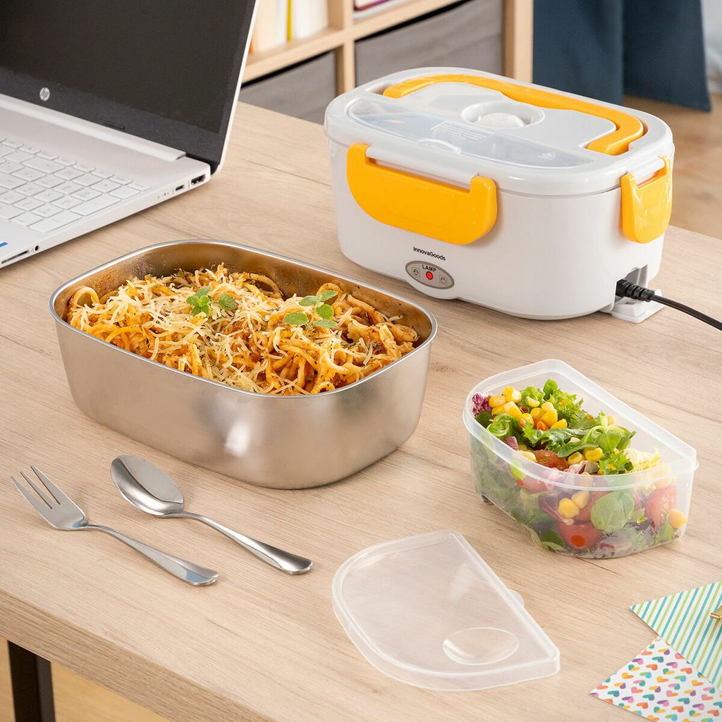 A portable electric lunch box with food and utensils on a wooden desk next to a laptop.