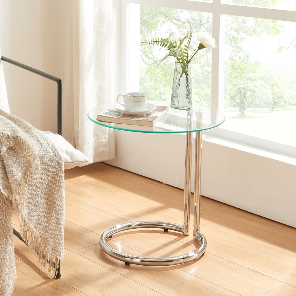 A glass-top side table with a chrome base sits next to a chair draped with a blanket, near a window with sheer curtains.