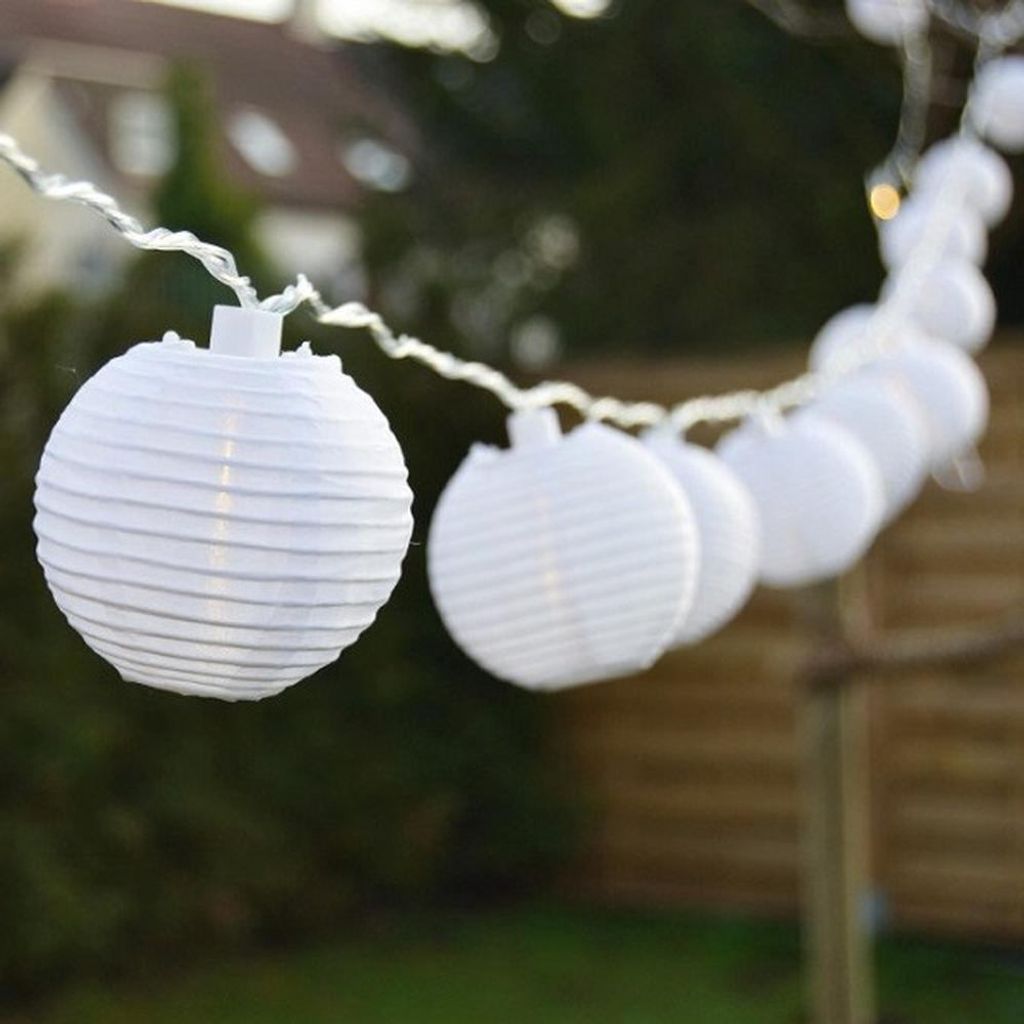 A string of white paper lanterns lit with warm white LEDs hangs outdoors, strung between two poles. The background shows greenery and a wooden fence.