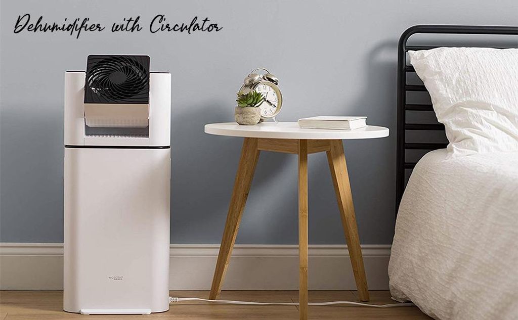 A white dehumidifier with a black fan is placed next to a bedside table with a book and a small plant on it, in a bedroom setting.
