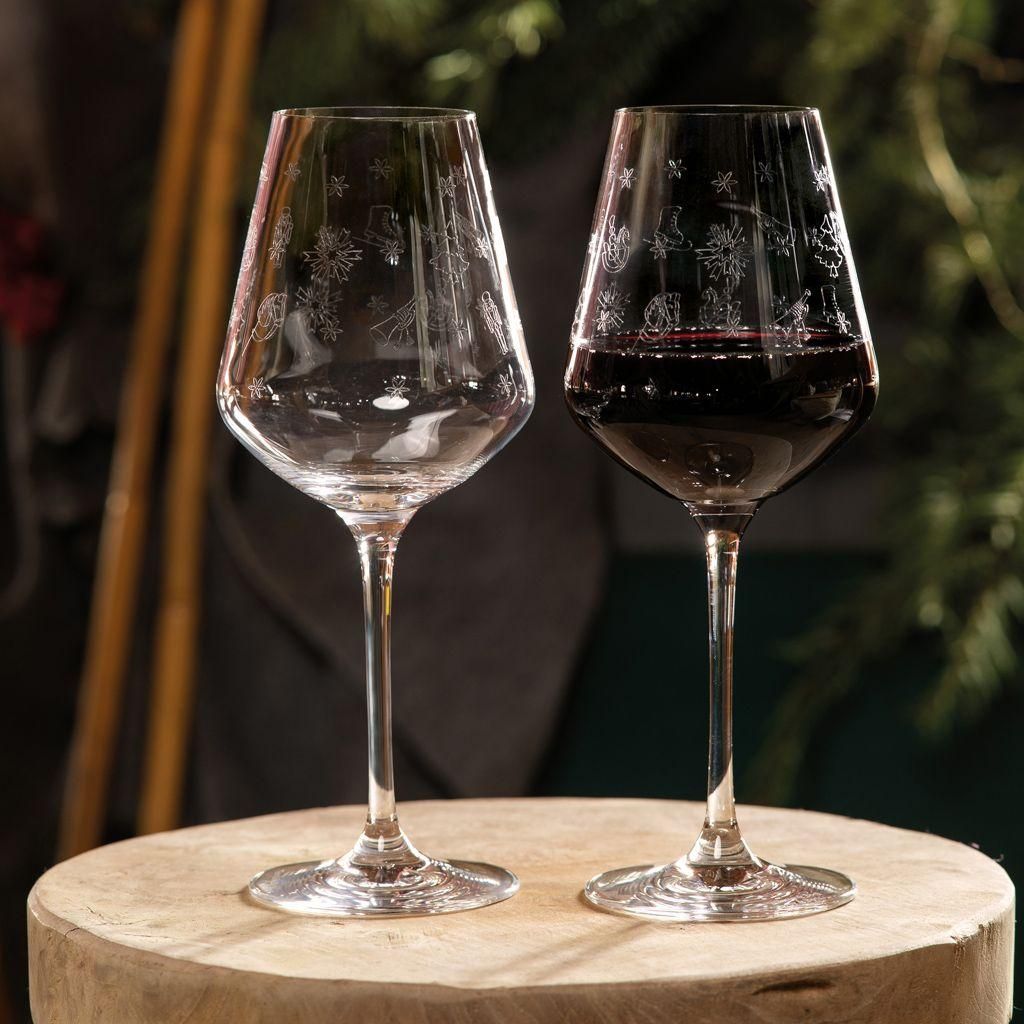 Two wine glasses, one empty and one filled with red wine, are placed on a wooden surface with a blurred background of greenery and wooden poles.