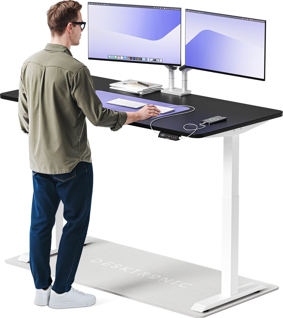 A man stands at a standing desk with two monitors, using a mouse. A mat is on the floor in front of the desk.