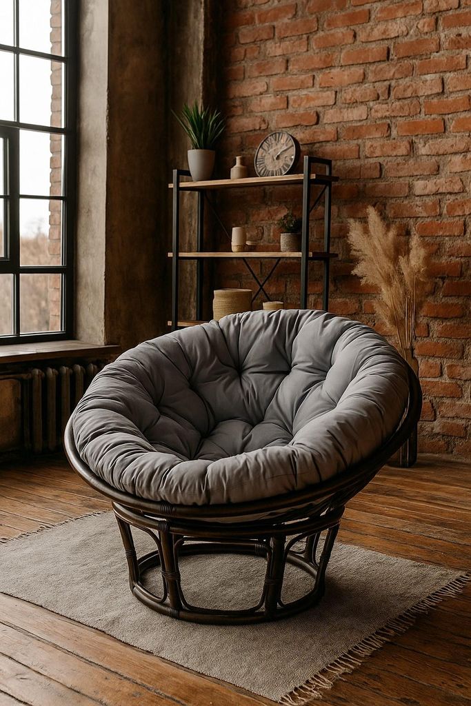 A papasan chair with a gray cushion sits on a rug in a room with exposed brick walls and a large window.