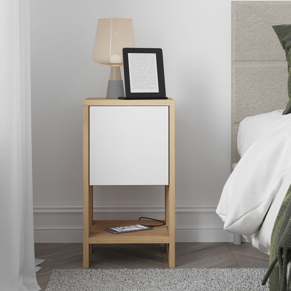 A bedside table with a drawer and an open shelf, placed next to a bed. A lamp and an e-reader are on top of the table, and a smartphone is on the shelf.