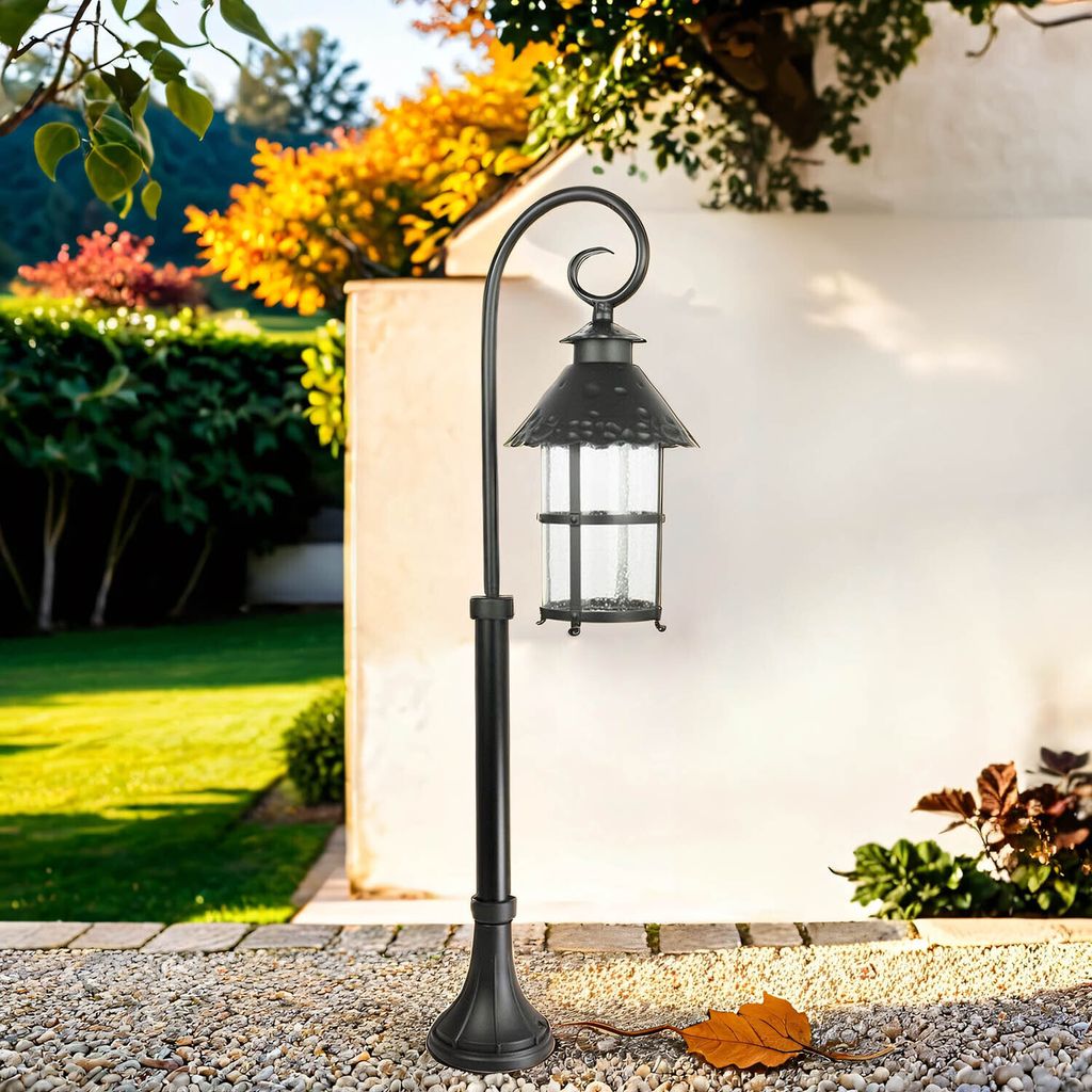 A black outdoor lantern on a post in a garden setting with autumn foliage.