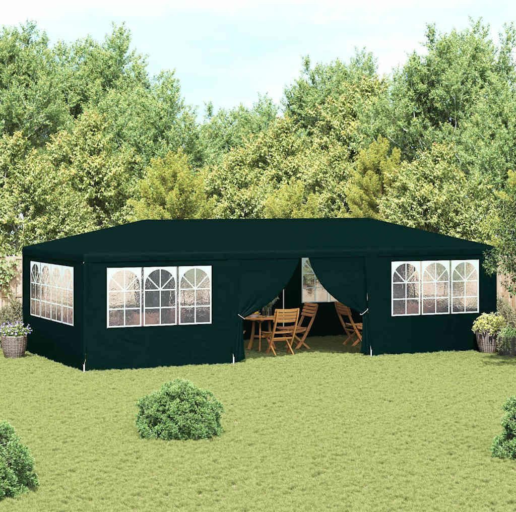 A dark green party tent set up in a grassy backyard with trees in the background. The tent has arched windows and an open entrance revealing tables and chairs inside.