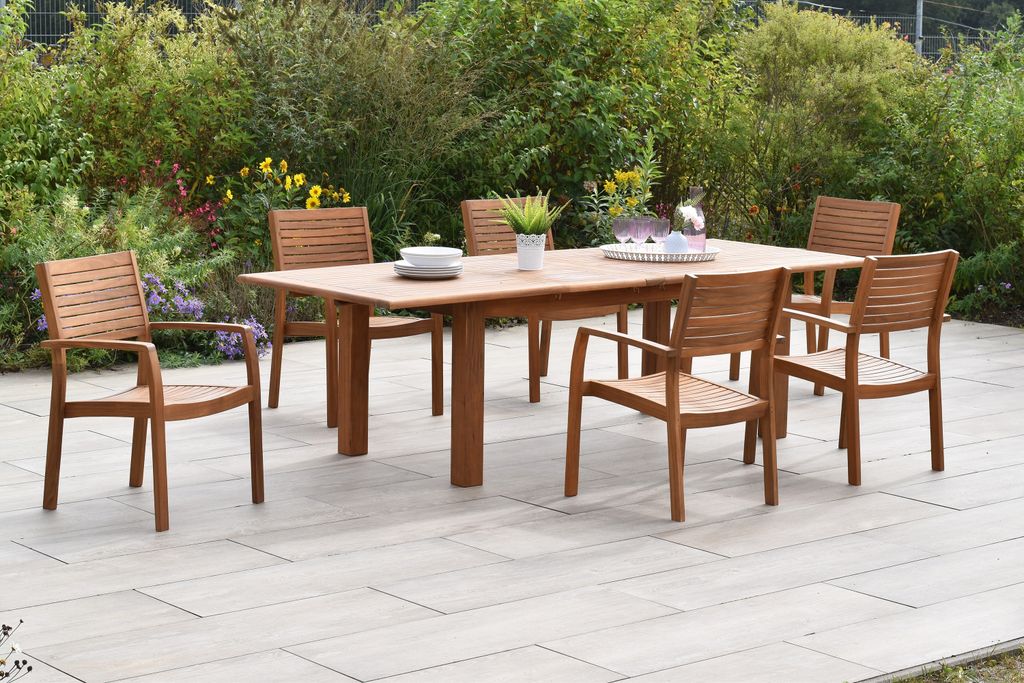A wooden dining set with six chairs and a table is placed on a patio surrounded by greenery and flowers.