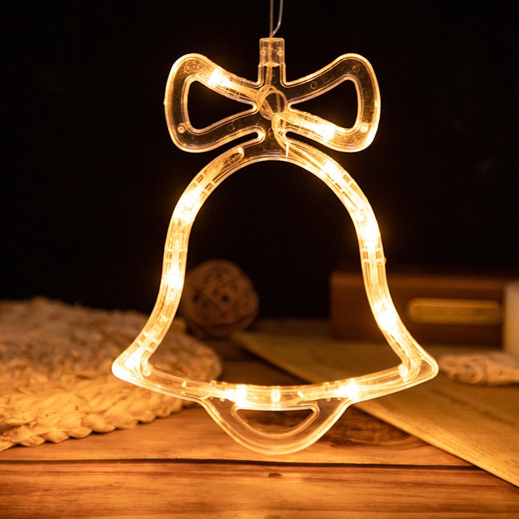 A close-up of a clear bell-shaped LED light with a bow on top, illuminated with warm yellow lights, hanging against a dark background. The bell is made of clear plastic and contains small LED lights. In the background, there are some decorative items like 