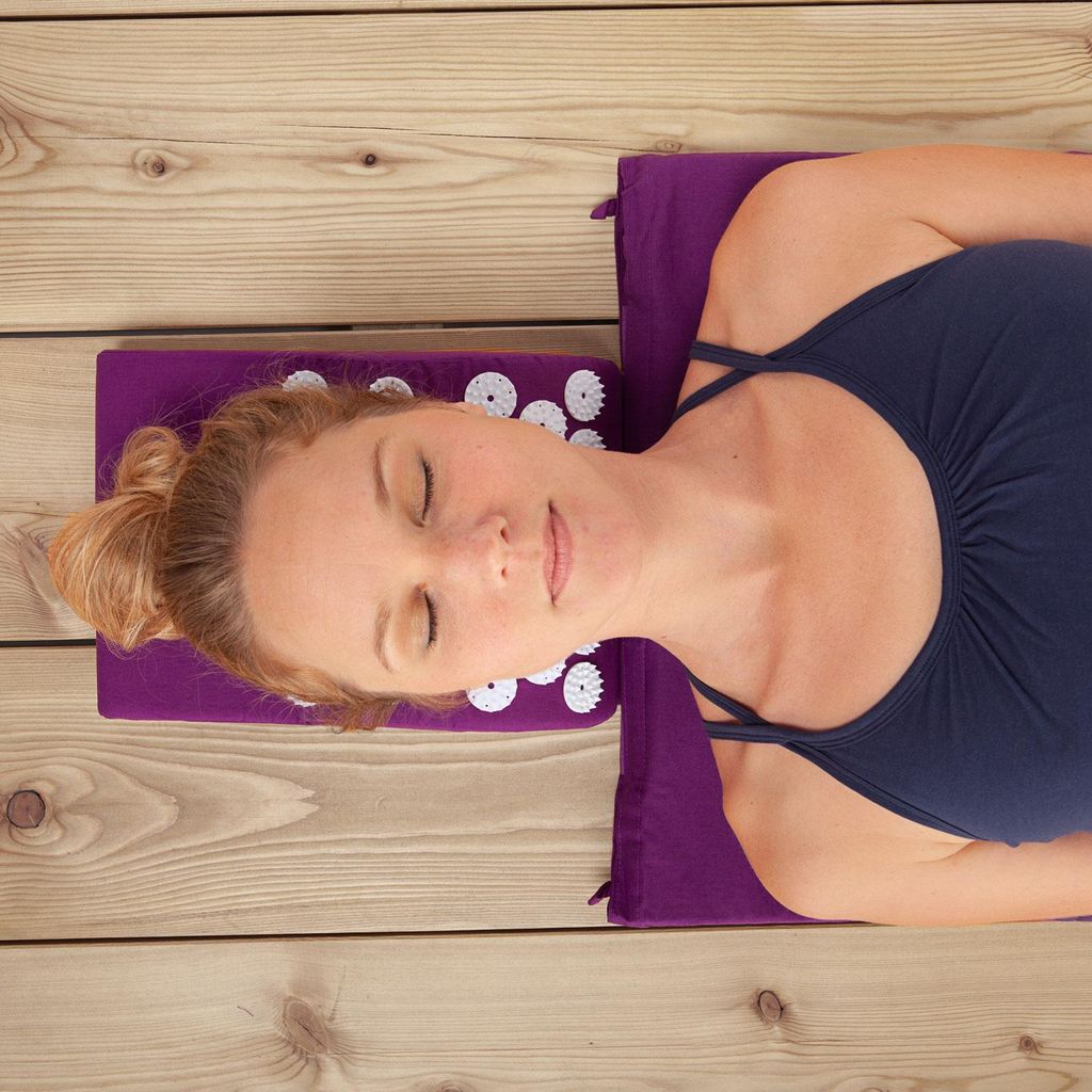 A woman is lying on her back on an acupressure mat and pillow set, which is placed on a wooden deck.